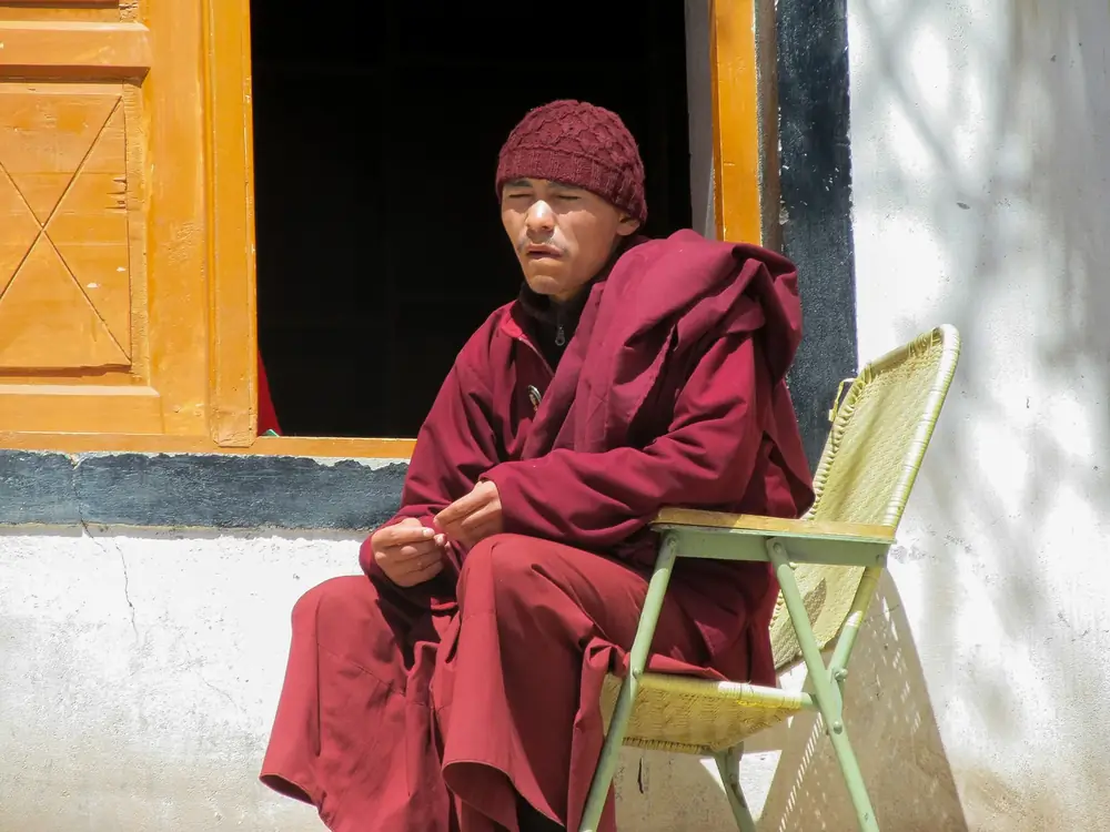 An elderly monk in maroon robes seated beside a sunlit wall